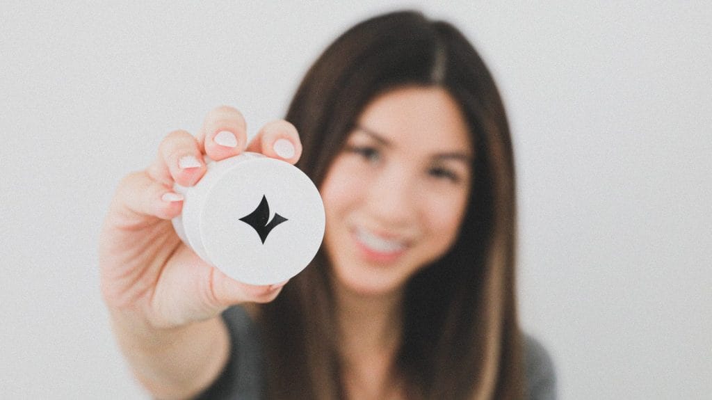 A person with brown hair holds a small white container featuring a black star-like logo towards the camera. The background is blurred.