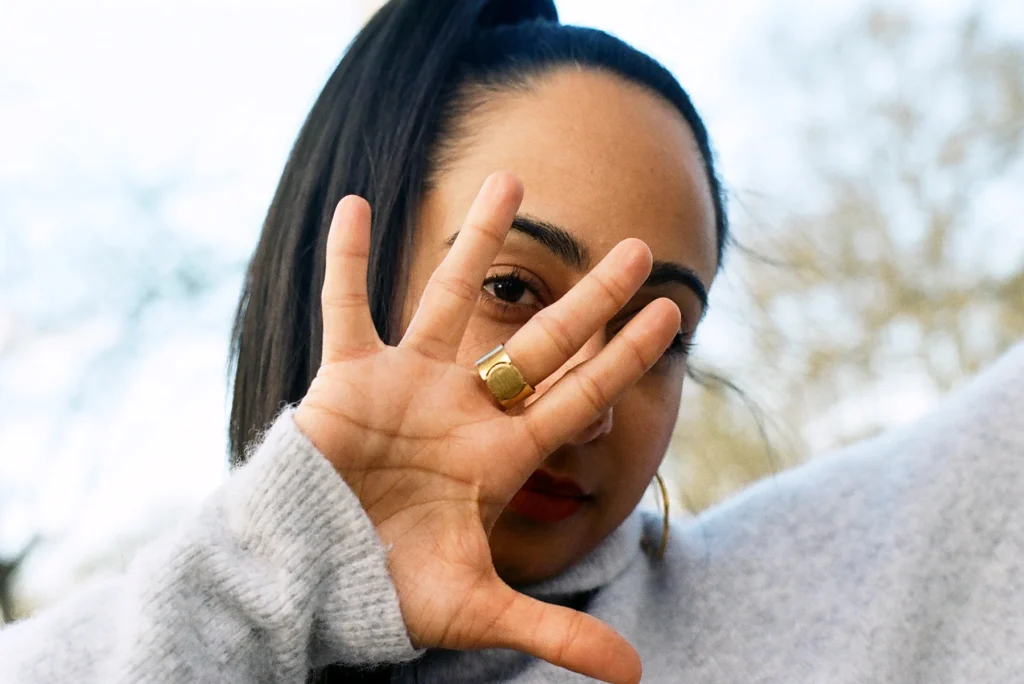 A person with long dark hair and a gold ring covers their face with a raised hand, standing outdoors.
