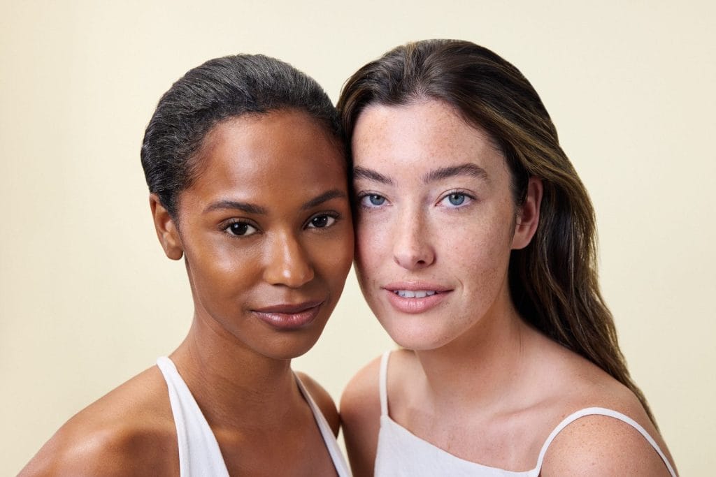 Two women in white tops stand closely together against a plain background, embodying the essence of safe and effective skincare, as they look directly at the camera with neutral expressions.