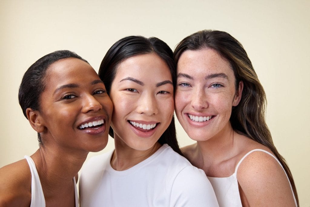 Three women are smiling and standing close together against a plain background, wearing light-colored tops that highlight their radiant glow, perhaps a testament to their favored skincare solution.
