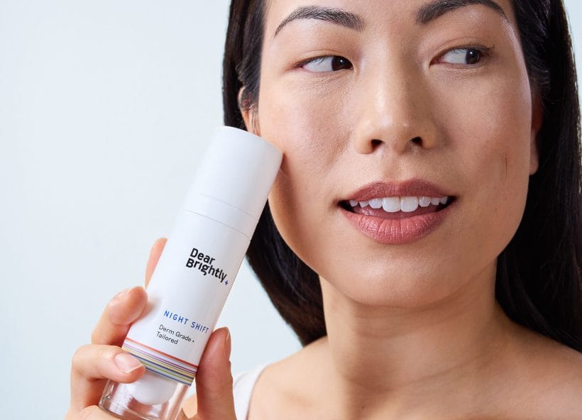 A person holds a bottle of Dear Brightly Night Shift cream, smiling slightly while applying the safe and effective skincare treatment to their face.