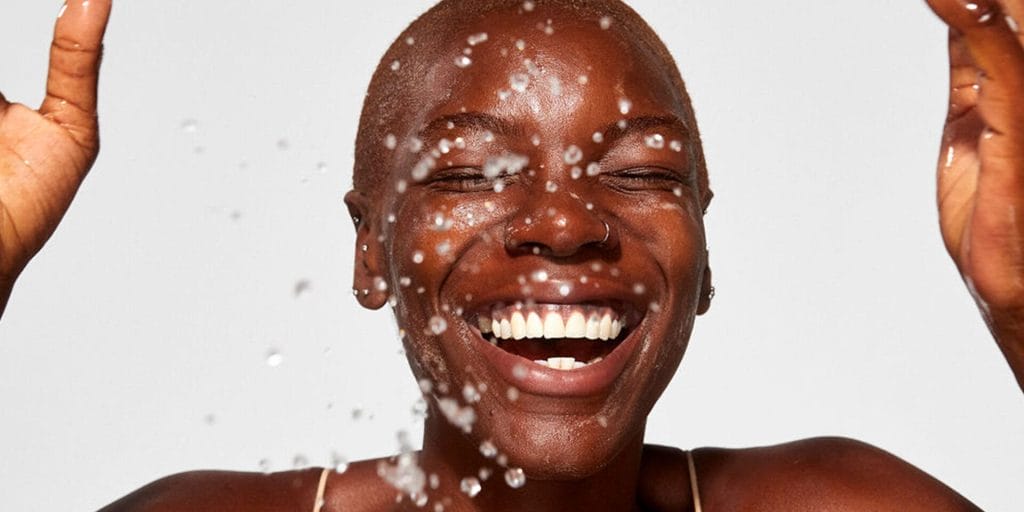 Person smiling with water droplets splashing around them against a plain background.