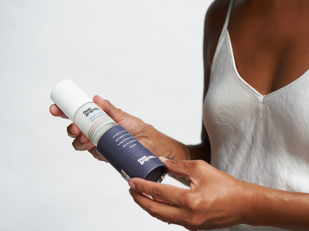 Person holding a bottle of deodorant spray with a dark label and white cap, wearing a white tank top.