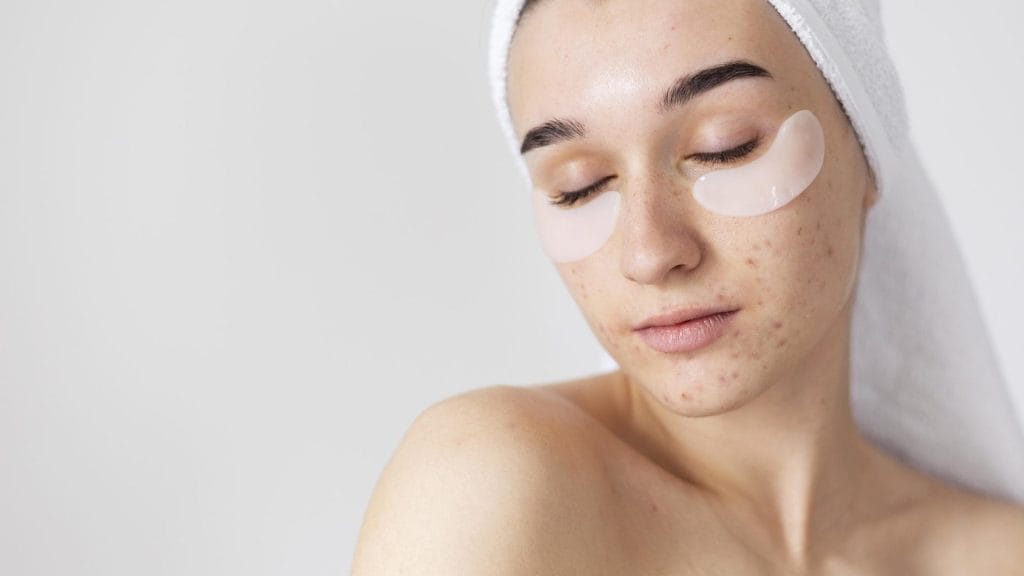 A person with a white towel wrapped around their head has closed eyes and under-eye patches on their face, likely benefiting from quality skincare products to address visible skin blemishes.